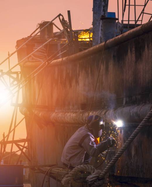 Side view of welder in protective workwear on platform is welding metal wall of the old fishing vessel in shipyard area at sunset time in vertical frame